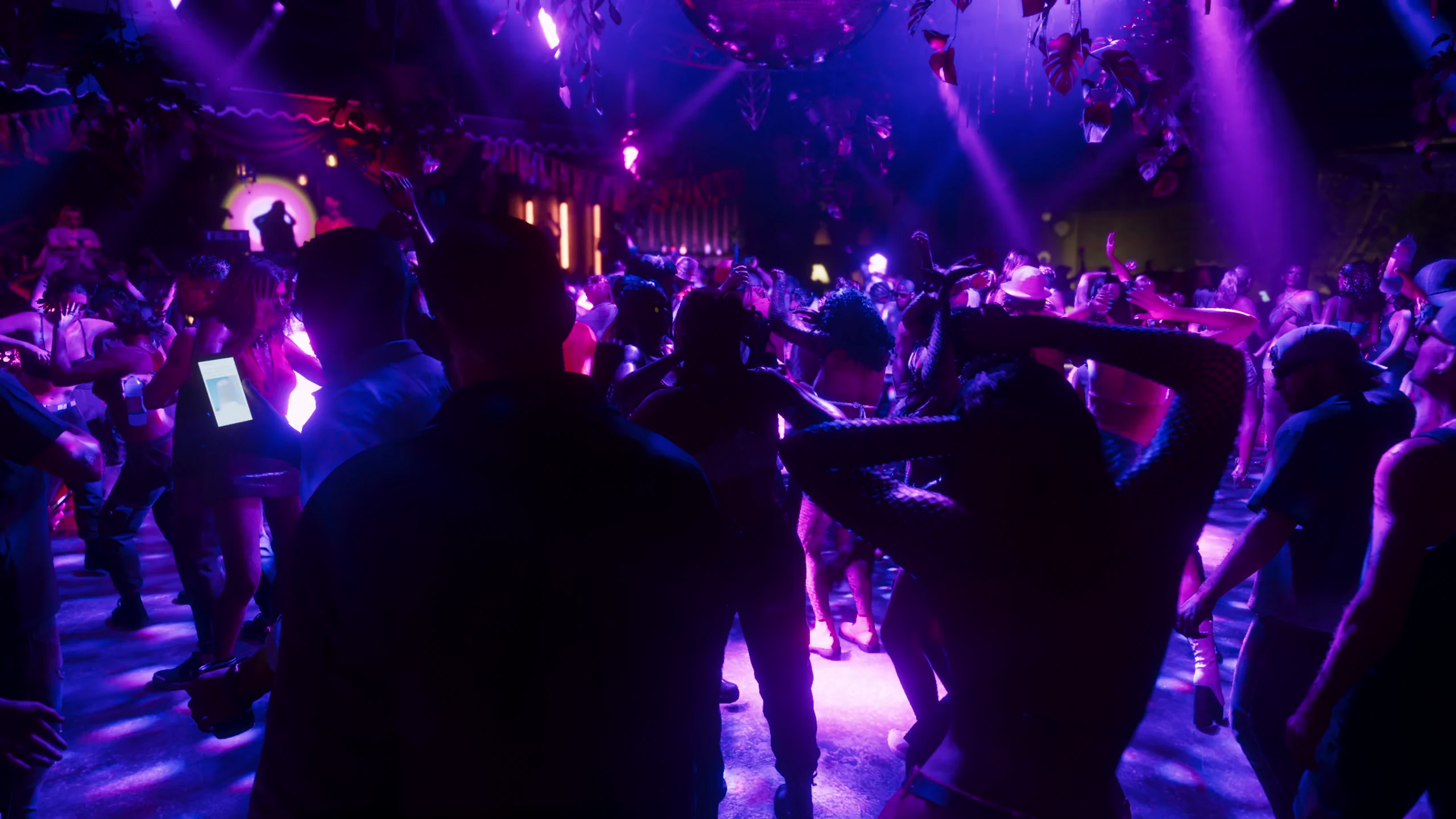 A packed nightclub floor under purple and pink lights, dozens of people dancing, seen from behind two figures moving into the crowd